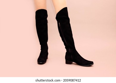 Black suede leather high long boots jackboots without heels, with flat sole and sexy Caucasian female legs on light powdery pink background. Studio shot photo, footwear concept.