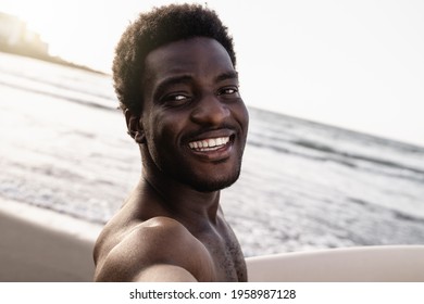 Black Surfer Man Doing A Selfie With Smartphone Holding Vintage Surf Board On The Beach At Summer Sunset - Focus On Face