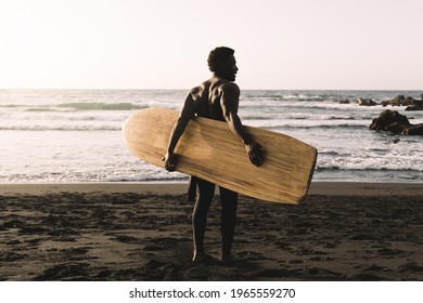 Black Surfer Man Holding Vintage Surf Board On The Beach At Sunset - Focus On Back