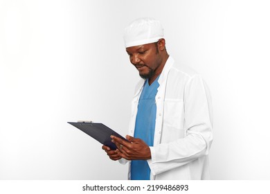 Black Surgeon Doctor Bearded Man In White Coat And Cap Looks To Medical Chart On Clipboard, Isolated On White Background Copy Space. Pensive Adult Black African American Practicing Surgeon Portrait