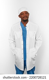 Black Surgeon Doctor Bearded Man In White Coat And White Cap With Hands In Pockets Isolated On White Background. Serious Adult Black African American Practicing Surgeon Portrait, Confident Look