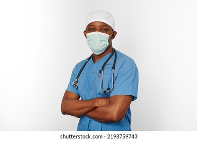 Black Surgeon Doctor Man In Blue Coat White Cap And Surgeon Mask With Crossed Arms Isolated On White Background. Focused Adult Black African American Practicing Surgeon Portrait, Confident Look