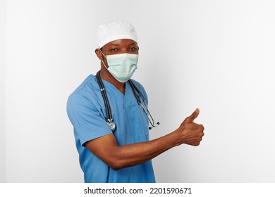 Black Surgeon Doctor Man In Blue Coat White Cap And Surgeon Mask With Thumbs Up Gesture Isolated On White Background. Focused Adult Black African American Practicing Surgeon Portrait, Confident Look