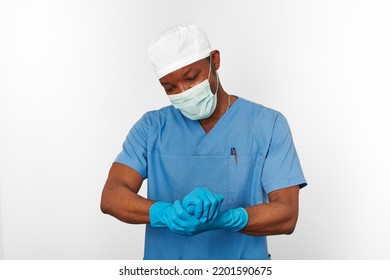 Black Surgeon Doctor Man In Blue Coat White Cap Surgeon Mask Sterilizes Blue Gloves With Aseptic Technique, Isolated On White Background. Adult Black African American Practicing Surgeon Portrait