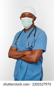 Black Surgeon Doctor Man In Blue Coat White Cap And Surgeon Mask With Crossed Arms Isolated On White Background. Focused Adult Black African American Practicing Surgeon Portrait, Confident Look