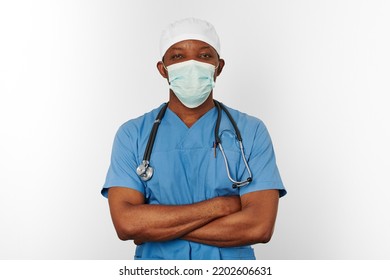 Black Surgeon Doctor Man In Blue Coat White Cap And Surgeon Mask With Crossed Arms Isolated On White Background. Focused Adult Black African American Practicing Surgeon Portrait, Confident Look