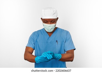 Black Surgeon Doctor Man In Blue Coat White Cap Surgeon Mask Sterilizes Blue Gloves With Aseptic Technique, Isolated On White Background. Adult Black African American Practicing Surgeon Portrait