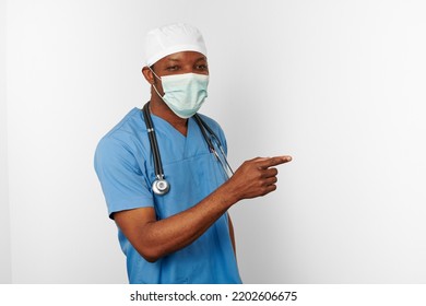 Black Surgeon Doctor Man In Blue Coat White Cap And Surgeon Mask Pointing Finger To Right Isolated On White Background. Focused Adult Black African American Practicing Surgeon Portrait, Confident Look