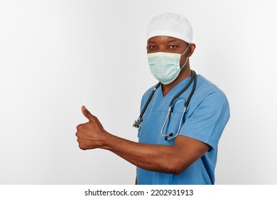 Black Surgeon Doctor Man In Blue Coat White Cap And Surgeon Mask With Thumbs Up Gesture Isolated On White Background. Focused Adult Black African American Practicing Surgeon Portrait, Confident Look