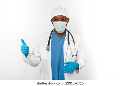 Black Surgeon Doctor Man In White Blue Coat Blue Gloves White Cap And Surgeon Mask Makes Thumbs Up Gesture Isolated On White Background. Friendly Black African American Practicing Surgeon Portrait