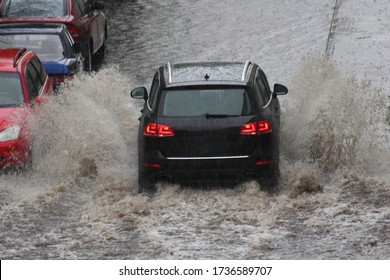 A black SUV car is driving fast with heavy spray in a deep puddle next to parked cars in heavy rain in the city on a spring day, the danger of hydraulic shock of engine, back view close up - Shutterstock ID 1736589707