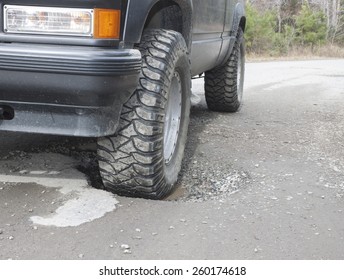 A Black SUV With A Front Wheel Sunk In A Water Filled Pothole On An Old Rural Asphalt/dirt Road