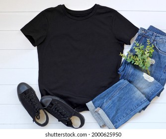 Black T Shirt Mock Up Flat Lay On White Wooden Background. Top Front View T Shirt And Copy Space. Mockup T Shirt With Denim, Eucalyptus And Black Shoes