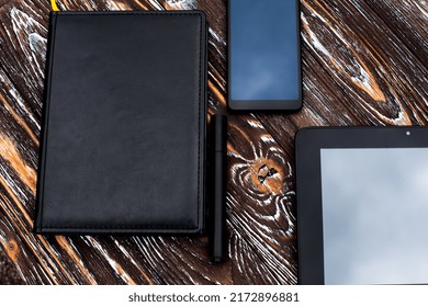A black tablet, a mobile phone, a notebook with a leather cover and a pen lie on a dark wooden textured surface.