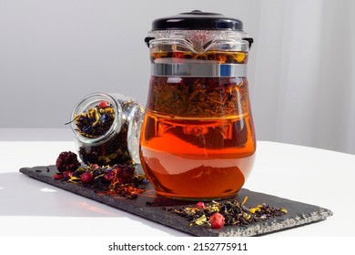 Black tea with herbs and berries in a transparent teapot on a white table. Jar with loose leaf tea. Hot brown drink. Advertising tea company, concept, presentation.