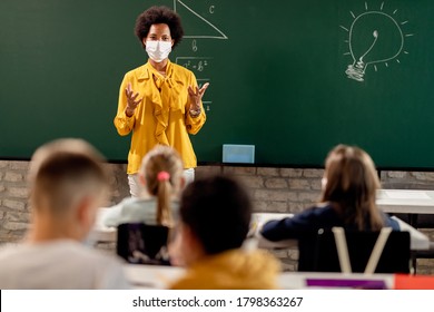 Black Teacher With Protective Face Mask Teaching Her Students On A Class At Elementary School.