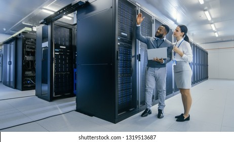 Black IT Technician with a Laptop Computer Gives a Tour to a Young Intern. They Talk in Data Center while Walking Next to Server Racks. Running Diagnostics or Doing Maintenance Work.