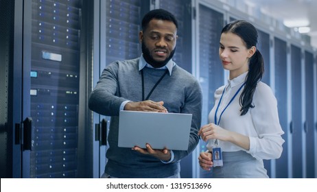 Black IT Technician with a Laptop Computer Gives a Tour to a Young Intern. They Talk in Data Center while Walking Next to Server Racks. Running Diagnostics or Doing Maintenance Work.