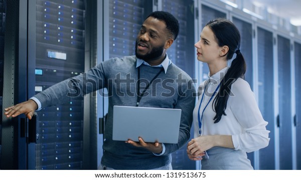 Black IT Technician with a Laptop Computer Gives a\
Tour to a Young Intern. They Talk in Data Center while Walking Next\
to Server Racks. Running Diagnostics or Doing Maintenance\
Work.