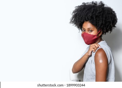 black teen girl shows the vaccine mark on her arm, wearing protective mask against covid-19, with a smile on her face, isolated on gray background