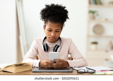 Black Teen Girl Using Mobile Phone Sitting At Laptop Computer Indoors. Home Education And Procrastination