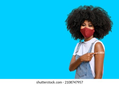 Black teen girl wearing face shield, points to the vaccine sticker on the square, you can see the smile in her eyes, isolated on blue background, covid-19