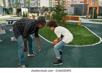 Black Teenage Boy Dribbling With Basketball Ball With His Father In Yard Of Modern District. Family Relationship And Spending Time Together. Fatherhood And Parenting. Entertainment, Hobby And Leisure