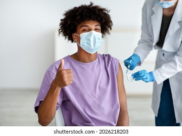 Black Teenager In Face Mask Making Thumb Up Gesture During Covid Vaccination, Approving Of Coronavirus Immunization At Clinic. Doctor Giving Antiviral Vaccine Injection To Afro Teen Patient