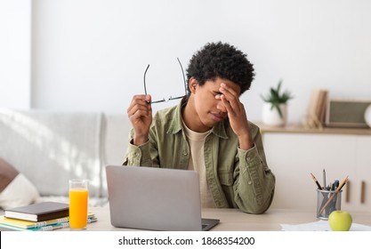 Black teenager feeling tired, rubbing irritated eyes, sitting at desk with laptop, exhausted from studying online. African American guy feeling fatigue from looking at screen, doing assignments on web