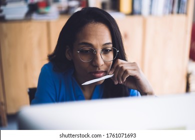 Black thoughtful serious busy female in eyeglasses working at project on computer and thinking about new ideas in office holding pen on chin