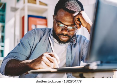 Black Thoughtful Serious Creative Author Taking Notes On Notebook For Article While Leaning With Head On Hand With Fatigue During Study On Laptop In Room