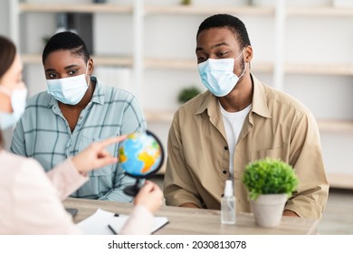 Black Tourists Couple And Travel Agent Lady Choosing Destination For Vacation Pointing At Globe Sitting In Tour Agency Office Indoors, Wearing Face Masks. Selective Focus