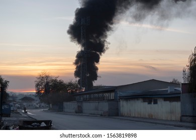 Black Toxic Smoke From Burning Garbage Rises Into The Sky And Pollutes The Environment. A Fire In A Landfill At Night In A Poor Area. An Unrecognizable Poor Man Drags A Cart In The Distance.