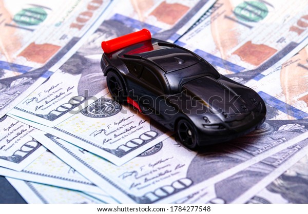 Black toy\
car on a heap of dollar bills on a white and black background. A\
sports car with a red spoiler, a wing on dollars in a close-up top\
and side view. July 2020. Los\
Angeles.USA