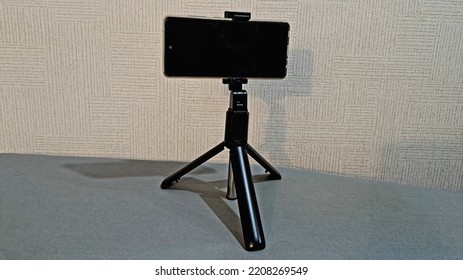 A Black Tripod With A Cell Phone Stands On A Table Near A White Wall In The Room