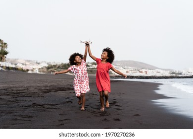 Black Twin Sisters Running On The Beach While Playing With Wood Toy Airplane - Focus On Faces