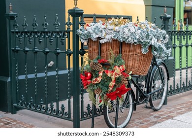 A black two wheel bicycle chained to a dark green wrought iron fence. The butcher's bike has a large wicker basket filled with greenery and Christmas flowers. There's a wreath with red bows on front.  - Powered by Shutterstock