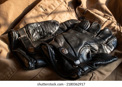 Black Vintage Leather Motorcycle Gloves. 1950's Short Biker Gloves With Leather Buckle Strap. Used, Worn And Weathered Motorbike Safety Gear. Stylish Retro Classic Placed On A Tan Colored Jacket