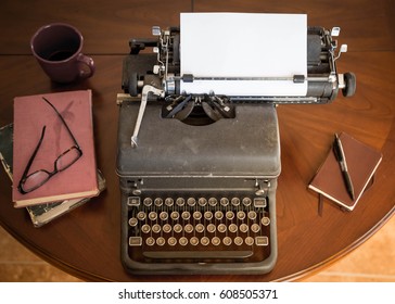 Black Vintage Typewriter Sitting On Brown Wooden Table Next To Brown Leather Notepad And Pen, And Old Books And Stylish Glasses. Any Author, Writer, Editor Or Journalist Can Type A Creative Story  