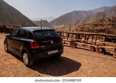 A black Volkswagen Polo car is parked in a rural area with a mountainous backdrop. In the background, there is a small outdoor eatery with wooden tables and benches. 