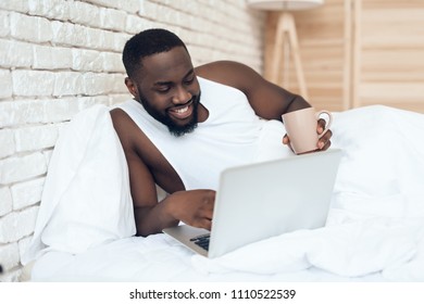 Black waking man drinks coffee in bed while working with laptop. Early morning. Pleasant awakening.