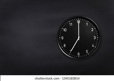 Black wall clock showing seven o'clock on black chalkboard background. Office clock showing 7am or 7pm on black texture