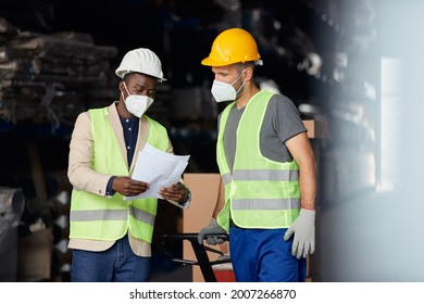 Black Warehouse Manager And Manual Worker Wearing Face Masks While Analyzing Paperwork At Storage Compartment. 