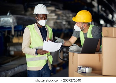 Black Warehouse Manager And Worker Communicating While Analyzing Reports At Distribution Warehouse. They Are Wearing Protective Face Masks Due To COVID-19 Pandemic.