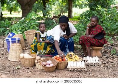 Black West African street market vendor displaying her homegrown products sitting on the edge of a village street with two of her daughters