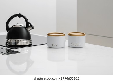 Black Whistle Kettle On Induction Cooktop Built In Glossy Kitchen Counter With Jars Next To It, Getting Ready To Make Tea With Sugar, White Furniture With Integrated Appliances