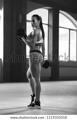 Black and white backview of athletic sporty woman posing with dumbbells in spacy gym with panoramic windows. Having strong, fit body with heatlthy tanned skin and muscles. Doing fitness exercises.