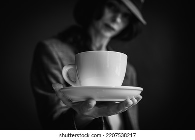 Black and white classic photo, girl in hat and gray jacket holding white cup with morning coffee and tea on gray background, close up view. Selective focus - Powered by Shutterstock