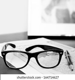 Black And White Close Up Still Life Of A Pair Of Reading Glasses And A Pen On An Orange Financial Newspaper On A Dark Wooden Writing Desk With A Family Photo Frame. Office Interior With No People.