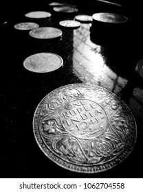Black and White closeup of a 1941 Indian British colonial era silver coin with intricate flower carvings, placed on dark black surface with sunlight reflections & array of coins further in background
